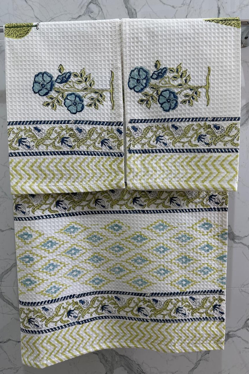 A set of three waffle-weave cotton towels hanging on a silver towel bar against a white marble wall. The towels feature a white base with a blue floral motif, olive green and navy blue geometric borders, and a central diamond pattern.