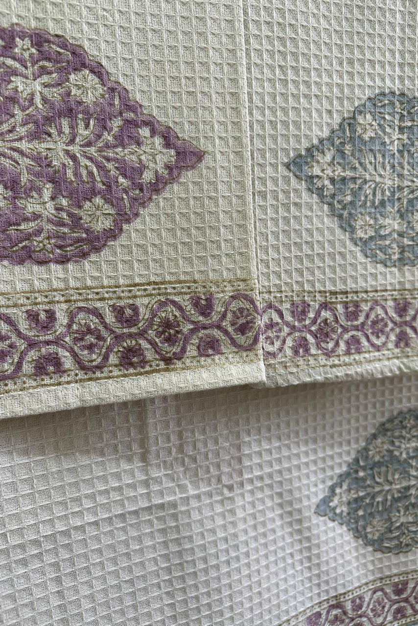 A close-up of a textured waffle-weave fabric, likely a cotton tablecloth or napkin, featuring a cream base with purple and dusty blue floral motifs and a decorative border.