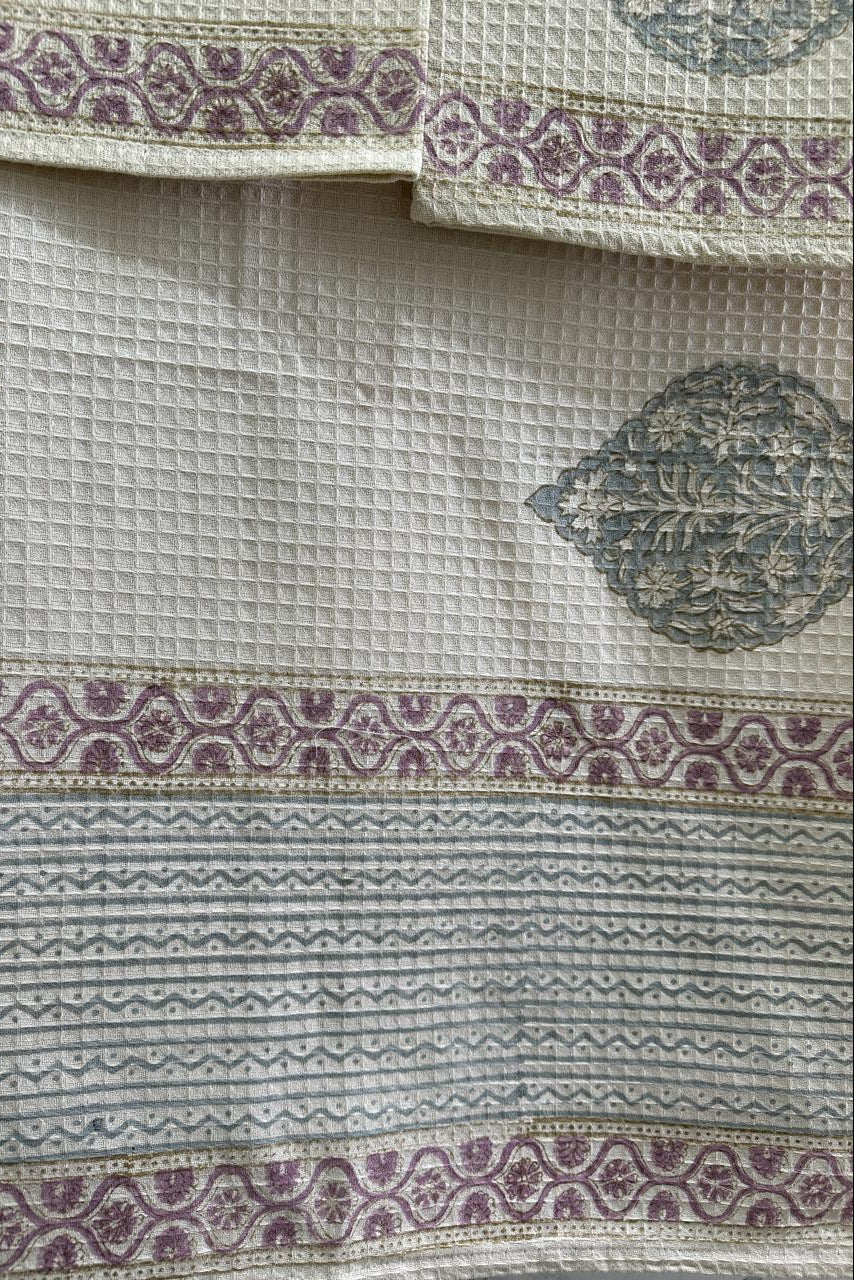 A close-up of a textured cream-colored fabric with woven geometric patterns and purple floral borders, showing part of a matching embroidered patch.