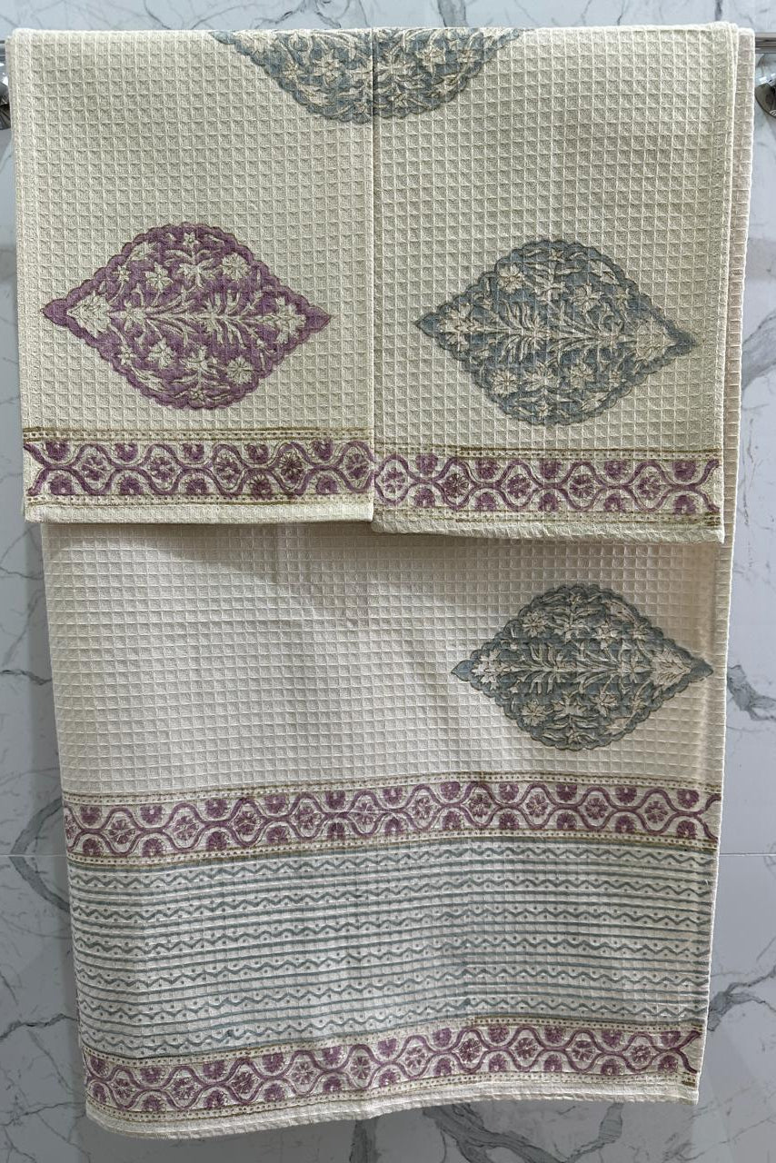 A set of two cream-colored waffle-weave towels hanging on a metal rack against a white marble-patterned wall. The towels feature a central wavy motif in dusty purple and gray, with a decorative patterned border in matching purple and gray.