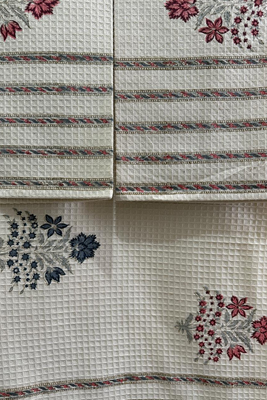 A close-up of a white waffle-weave fabric textile, likely a tablecloth or napkin set, featuring embroidered floral motifs in red, blue, and pink with decorative borders.