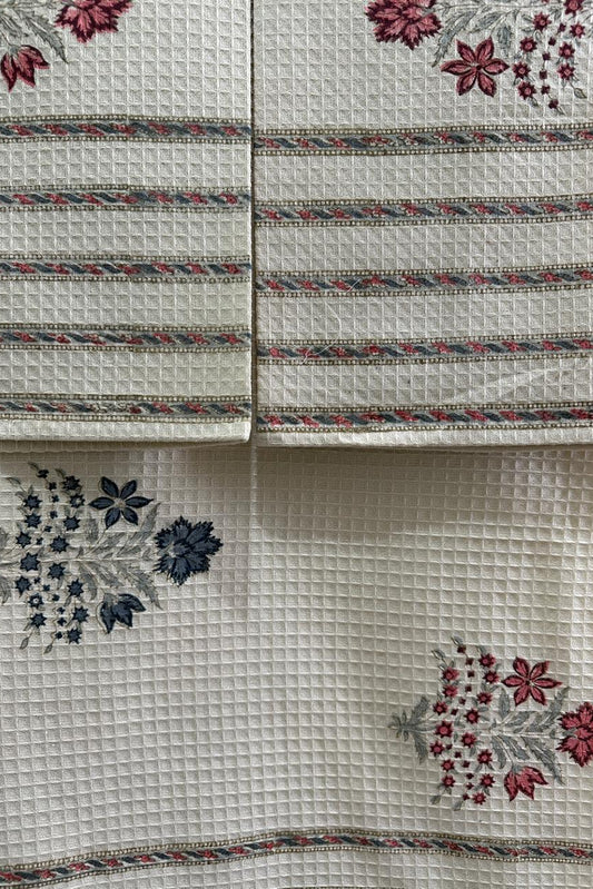 A close-up of a white waffle-weave fabric textile, likely a tablecloth or napkin set, featuring embroidered floral motifs in red, blue, and pink with decorative borders.