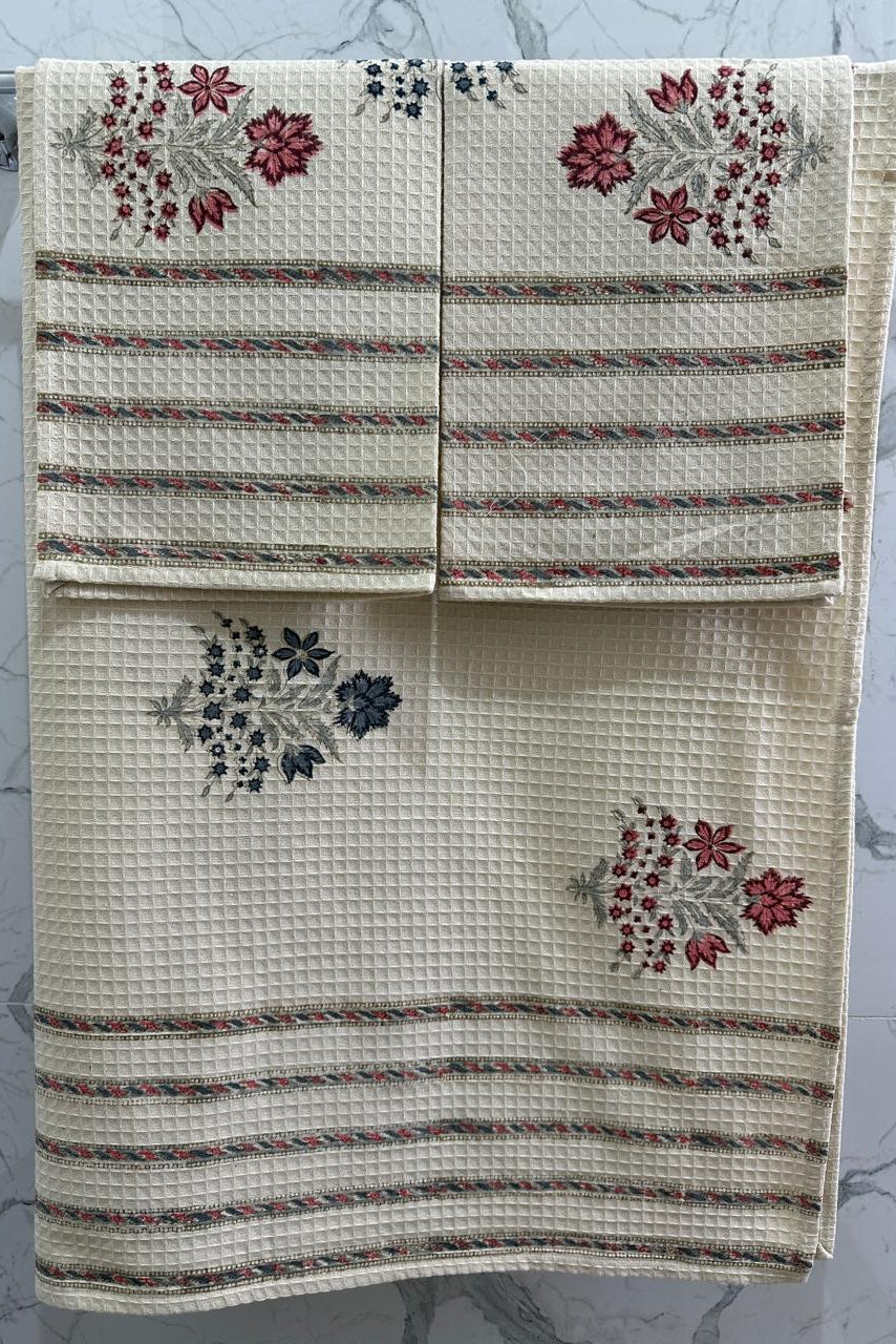 Three cream-colored waffle-weave kitchen towels with striped and floral patterns, hanging on a silver metal rack against a white marble-patterned wall.