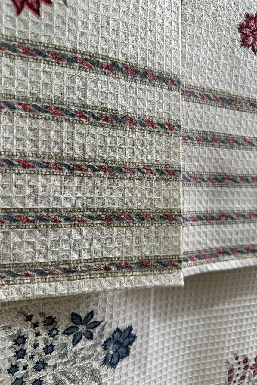 Close-up of a textured cream fabric with woven horizontal stripes in red, blue, and gray, featuring floral embroidery in matching colors.