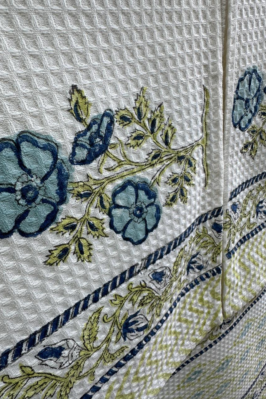 Close-up of a white waffle-weave textile with embroidered blue flowers, green leaves, and a decorative border pattern.