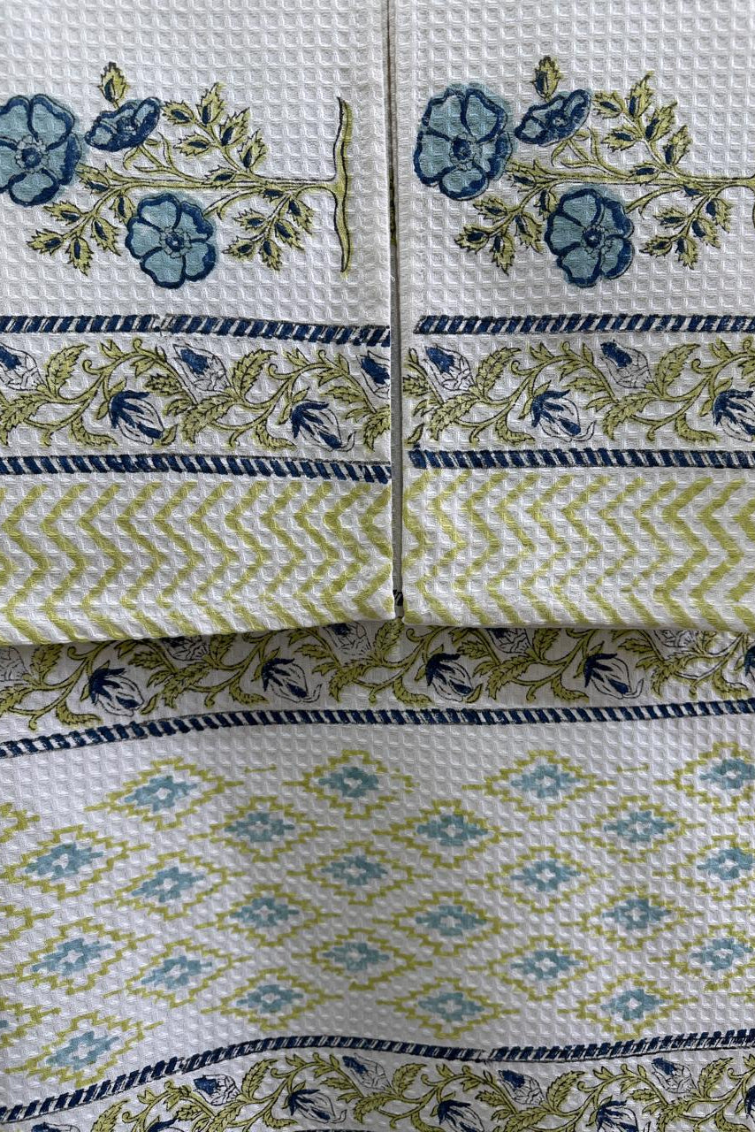 A close-up of a folded white waffle-weave textile, likely a towel or napkin, featuring blue floral motifs, olive green and navy decorative borders, and a repeating blue and green diamond pattern.