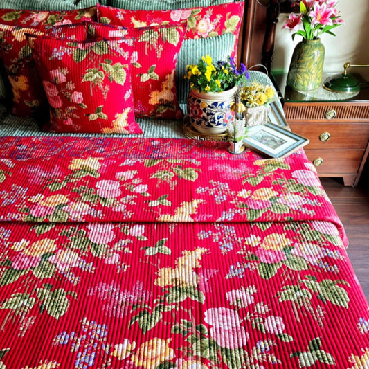 Bed with red floral bedding and pillows, side table with flowers and lamp in a bedroom setting.