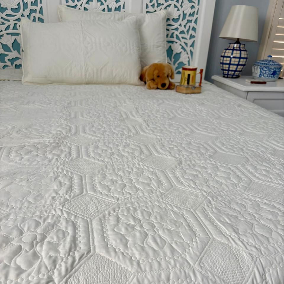 Textured white quilt on a bed with decorative pillows and a lamp in the background.