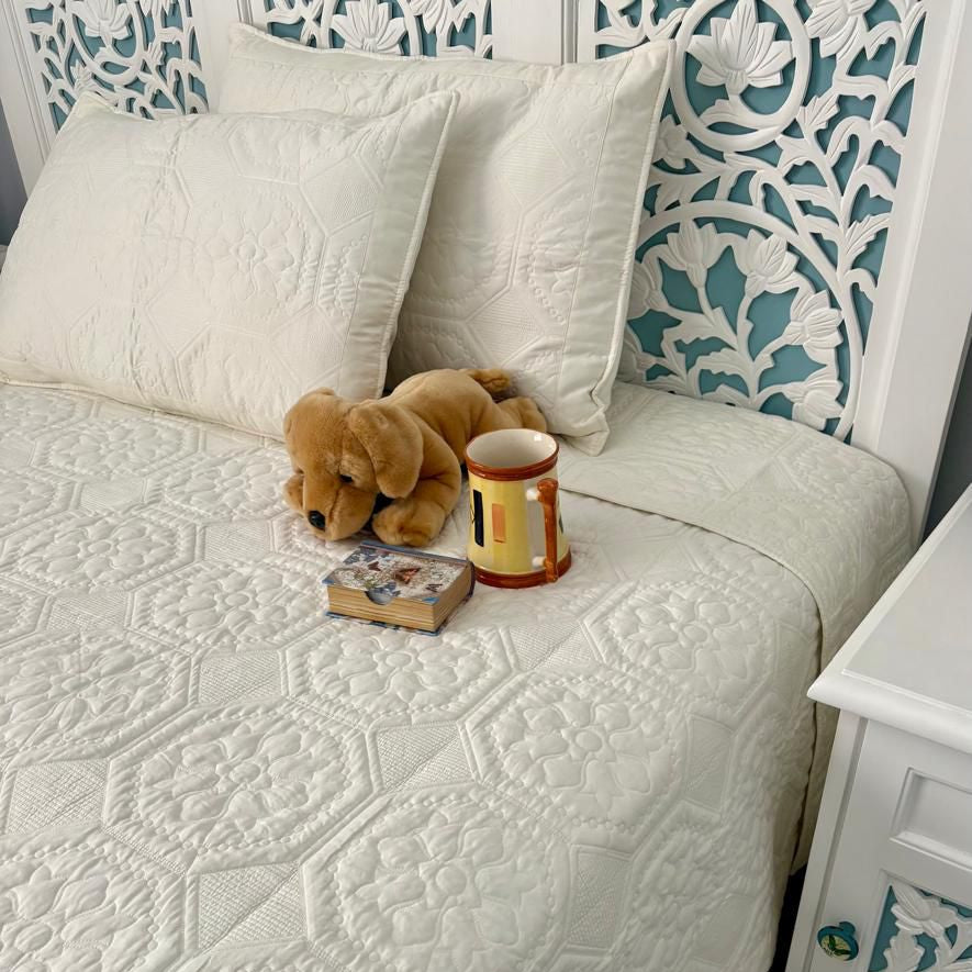 Bed with textured white cover, teddy bear, and book in a room with floral-patterned wallpaper.