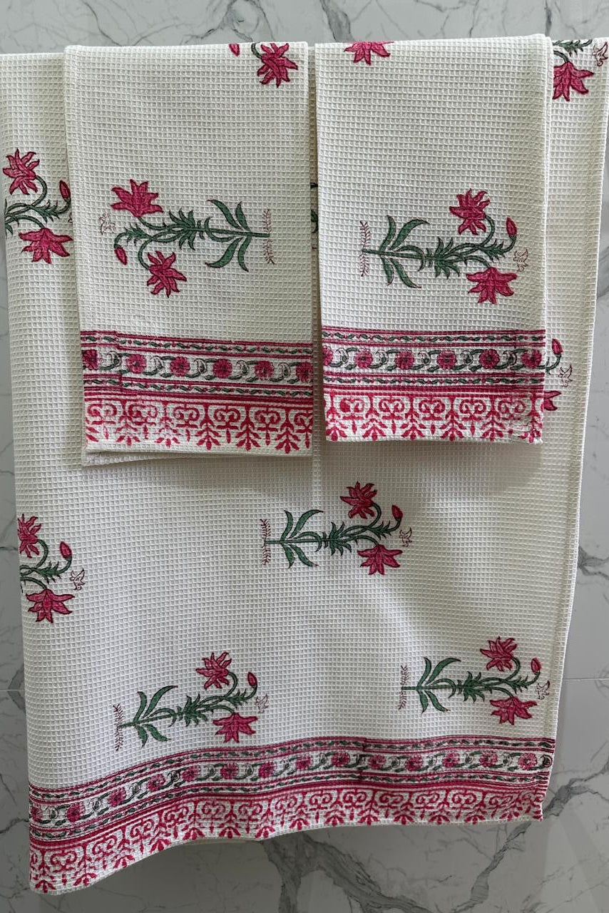 A set of three white waffle-weave kitchen towels with red floral motifs and green stems, hanging on a metal rack against a gray marble-patterned wall.