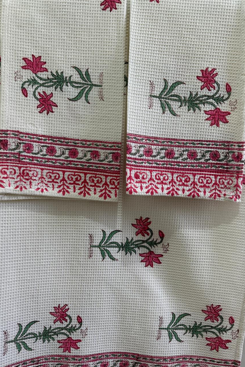 A close-up of a white or off-white woven textile, likely a napkin or small table linen, featuring pink floral motifs with green stems and leaves, and a decorative pink and green border.