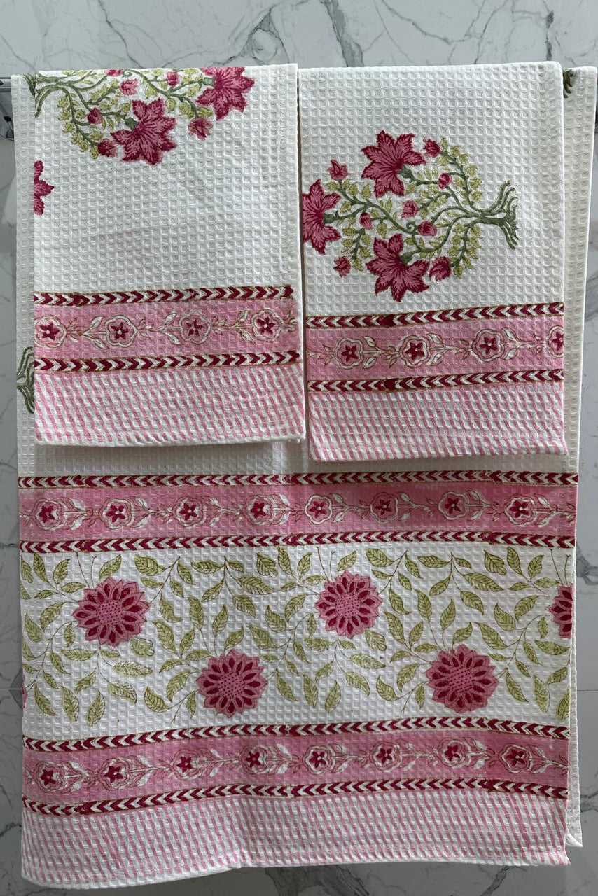 A set of three white waffle-weave kitchen towels with pink floral and green leaf patterns, hanging on a silver metal rack against a light gray marble-patterned wall.
