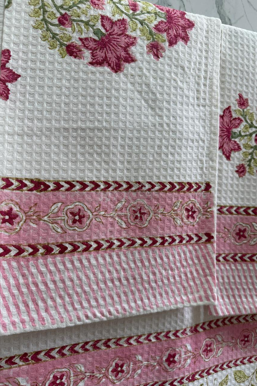 A close-up of a white waffle-weave kitchen towel with pink and green floral embroidery and decorative pink borders.