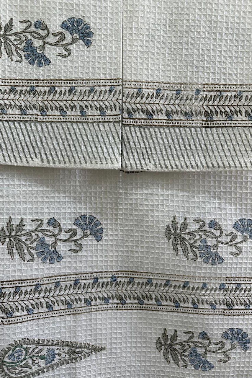 A close-up of a white textured fabric, likely cotton muslin or waffle-weave, featuring hand-block printed floral motifs and horizontal decorative borders in blue and gray.