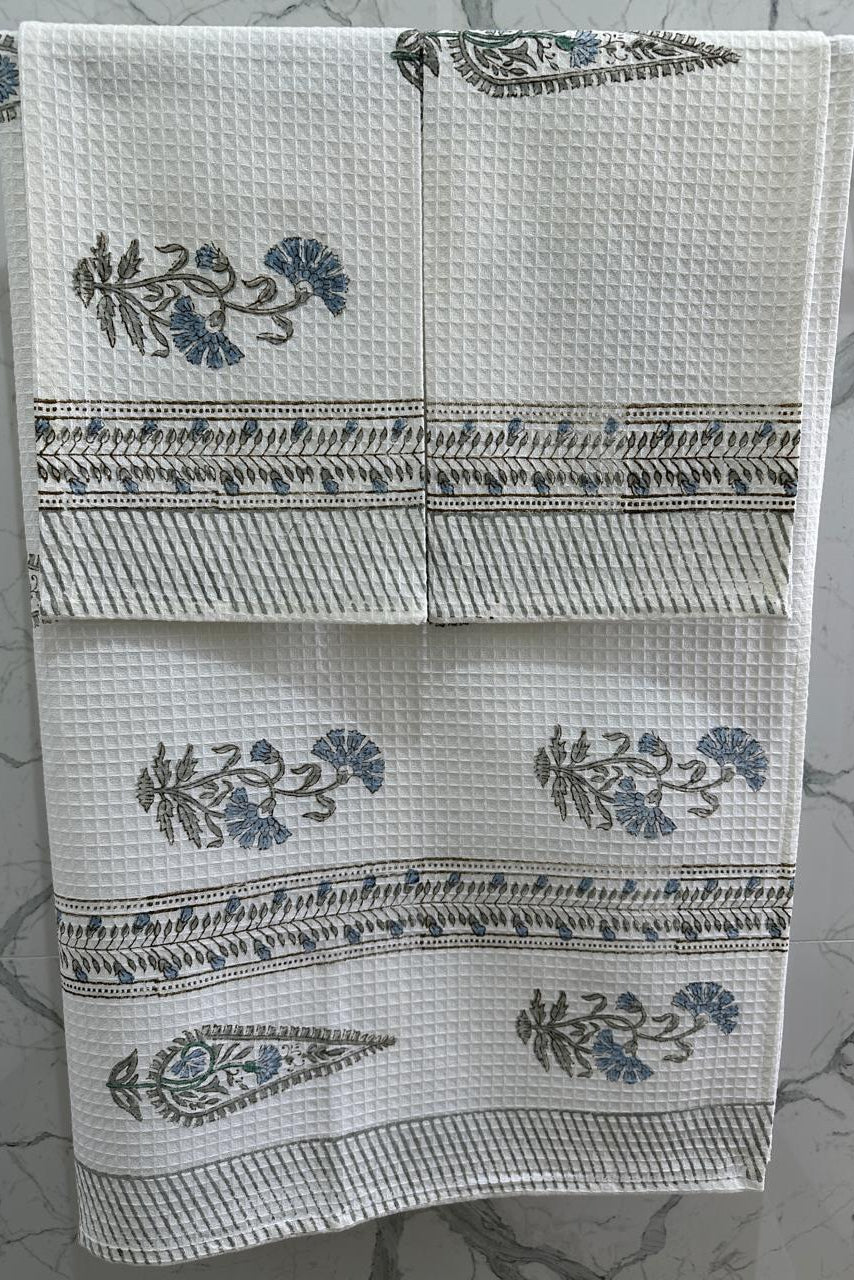 A set of three white waffle-weave kitchen towels hanging on a metal rack against a gray marble-patterned wall. Each towel features a blue floral motif and a decorative border.