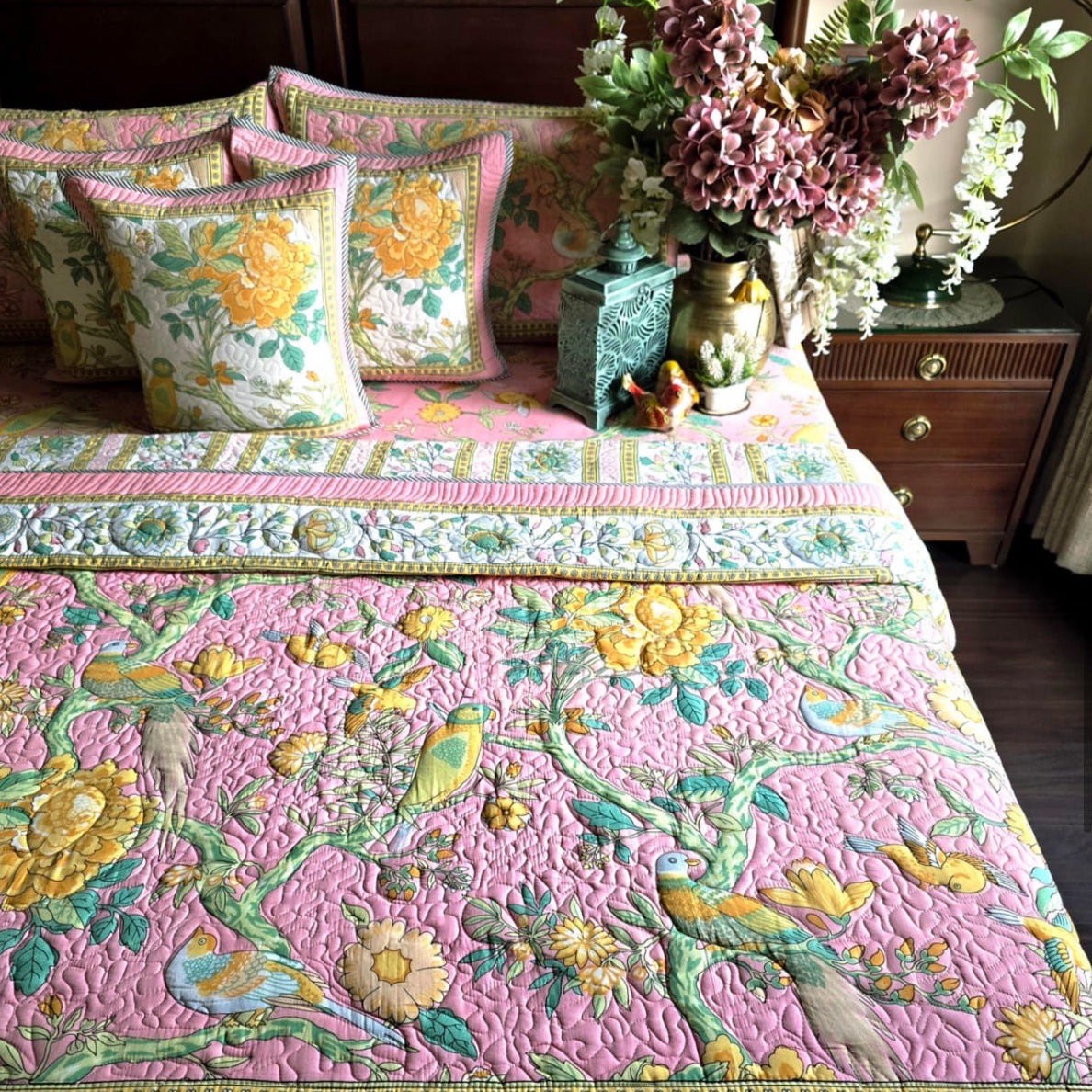 Bed with floral quilt and pillows, decorated with flowers and a vase on a nightstand.