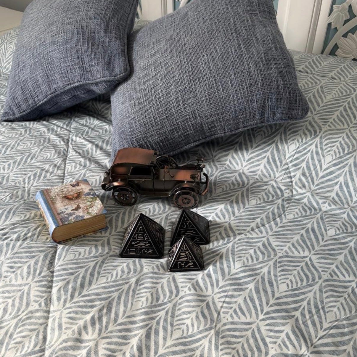 Small model car and black triangular objects on a bed with gray pillows and patterned bedding.